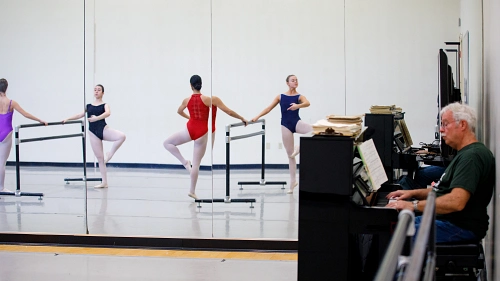 Ballet dancers practice at the barre in a studio while an older man plays piano in the foreground.