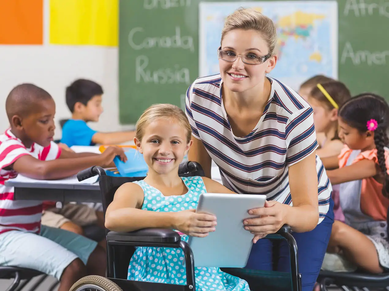 special education teacher with student in wheelchair looking at camera with students in the background working at a table